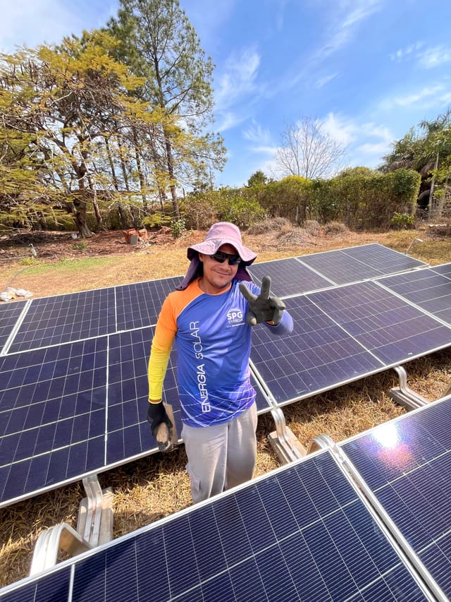 Instalador com uniforme da SPG Energia acenando para a câmera entre fileiras de placas solares de uma usina em solo