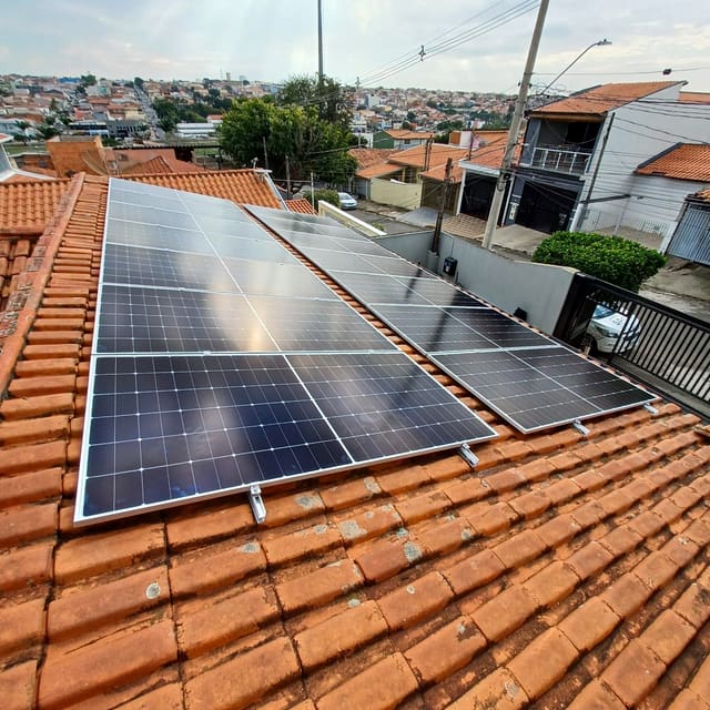 Placas solares instaladas sobre telhado de telha cerâmica de uma casa, com o bairro ao fundo