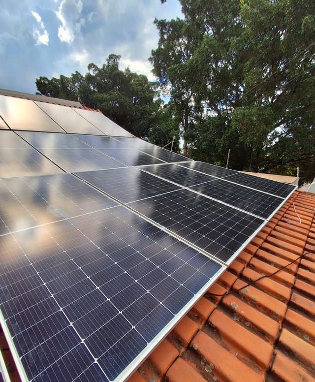 Fileira de placas solares fixadas sobre telhado de telha cerâmica, com árvores ao fundo