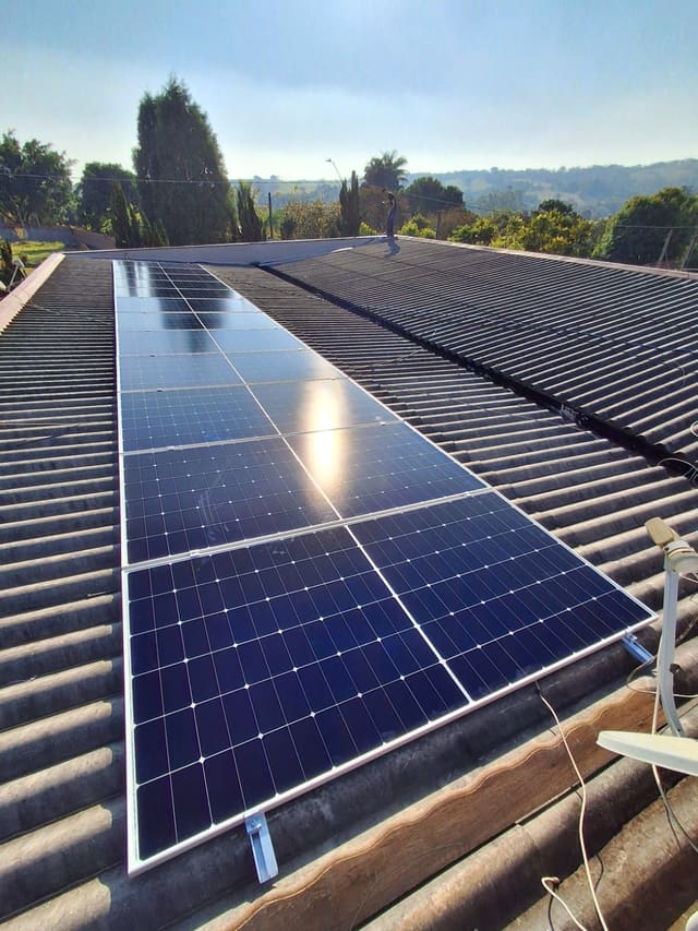 Placas solares em telhado de fibrocimento ondulado, com árvores e morros ao fundo