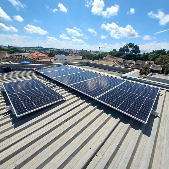 Placas solares fixadas em telhado metálico, com céu azul e o bairro ao fundo